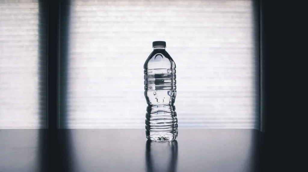 A clear reusable bottle filled with water beside a glass on a bright table